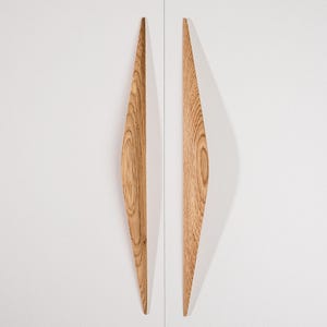 May include: Two vertical cabinet handles made of light brown oak wood with a visible natural grain pattern. The handles have a long, slender, tapered shape that is wider in the center and narrows toward the top and bottom ends, mounted on a white surface.