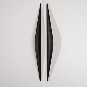 May include: Two elongated, vertical cabinet handles made of black wood with a visible grain texture. The handles feature a tapered, spindle-like shape that is thicker in the centre and narrows towards both ends, mounted on a white surface.