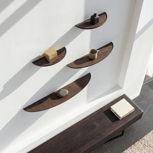Puede incluir: Cuatro estantes de pared de madera oscura en forma de media luna montados en disposición escalonada. Los estantes sostienen un pequeño jarrón negro, una taza de cerámica beige, un cubo dorado y una piedra gris lisa. Un banco largo de madera oscura.