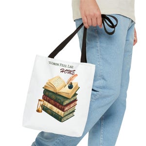 Op de afbeelding: Een witte tote bag met zwarte hengsels voorzien van een afbeelding van een stapel vintage boeken, een open boek met een ganzenveer en inktpot, en een zandloper. De tekst "WORDS FEEL LIKE HOME" is in zwart en rood boven de boeken gedrukt.