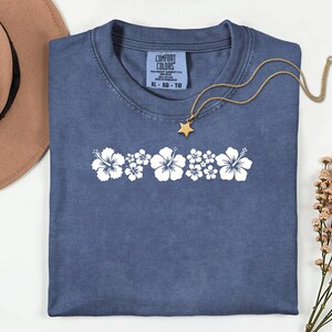 May include: A blue short-sleeved t-shirt featuring a horizontal row of white hibiscus flower graphics across the chest. The shirt has a Comfort Colors label at the neck with size XL, and a gold-toned necklace with a star charm rests on top.