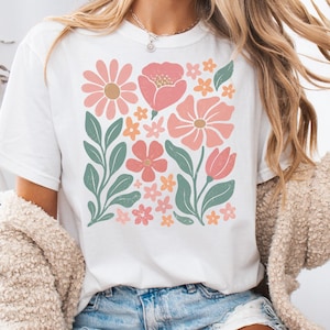 May include: A white short-sleeve t-shirt featuring a central graphic print of various stylized flowers and leaves in shades of pink, muted orange, and sage green. The floral design is arranged in a rectangular block pattern on the front chest.