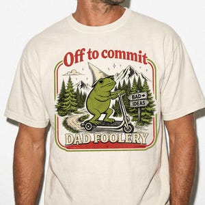 May include: A cream-coloured t-shirt featuring a graphic of a green frog wearing a pointed hat and riding a scooter. The text reads Off to commit DAD FOOLERY, with a sign pointing toward BAD IDEAS, set against a background of pine trees and mountains.