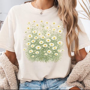May include: A cream-colored short-sleeve t-shirt featuring a central graphic print of a dense field of white daisies with yellow centers and green stems. The shirt has a crew neckline and a relaxed fit, showcasing a botanical floral illustration.