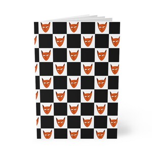 May include: A rectangular notebook featuring a repeating pattern of orange fox faces with white accents on a black and white chequered background. The cover displays multiple stylised fox heads arranged in a grid, alternating with black squares.