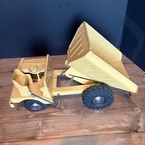 May include: A yellow metal toy dump truck featuring a steering wheel, an open driver seat, and a large tilting cargo bed. The vehicle is equipped with four black rubber tires with deep treads and silver-colored hubcaps on the wheels.