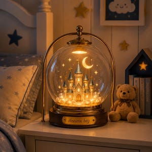 May include: A spherical glass lamp featuring an illuminated miniature castle with blue-roofed towers inside. The lamp has a gold-toned metal frame and a dark wooden base with a gold plaque engraved with the text "dream big" in cursive script.