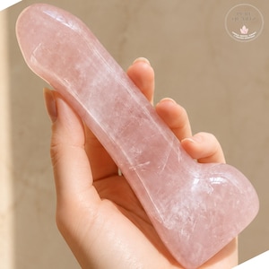 Peut inclure: Un bâton en quartz rose poli et lisse de forme phallique, doté d'une extrémité arrondie et d'une base évasée. La pierre présente une teinte rose pâle translucide avec des inclusions naturelles et une finition brillante. Texte Pure Quartz.