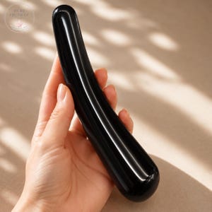 May include: A smooth, polished black obsidian wand with a curved, cylindrical shape and a rounded tip. The surface is highly reflective, showing light highlights along its length. The object is solid, opaque, and dark in color throughout.