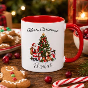 May include: A white ceramic mug with a red interior and handle, featuring a printed illustration of Santa Claus, a decorated Christmas tree, a snowman, and a reindeer. The text Merry Christmas and the name Elizabeth are printed in black script.