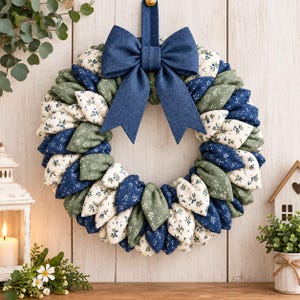 Op de afbeelding: Een stoffen krans met een cirkelvormige opstelling van opgevulde, bloembladachtige stoffen stukjes in marineblauw, saliegroen en crème met kleine bloemmotieven. Een grote, effen marineblauwe stoffen strik is aan de bovenzijde bevestigd.