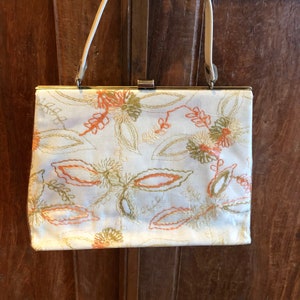 K&ouml;nnte beinhalten: Eine wei&szlig;e Handtasche mit einem floralen Stickmuster in Orange-, Gr&uuml;n- und Goldt&ouml;nen. Die Tasche hat einen Verschluss oben und einen einzigen Trageriemen.