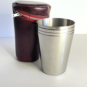 Könnte beinhalten: Ein Edelstahlbecher mit drei horizontalen Linien am oberen Rand, neben einem weinroten Lederetui mit rotem Reißverschluss. Das Etui ist zylindrisch und hat einen silbernen Reißverschluss. Der Becher ist leicht konisch.