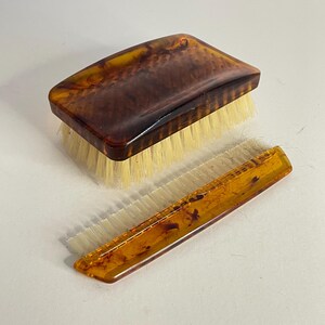 May include: A vintage grooming set comprising a rectangular brush and a comb. The brush and comb display a tortoiseshell pattern in brown and yellow tones. The brush is rectangular with white bristles.