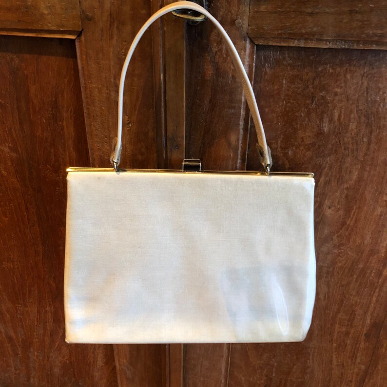 K&ouml;nnte beinhalten: Eine wei&szlig;e Clutch mit goldenem Rahmen und einem braunen Ledergriff.