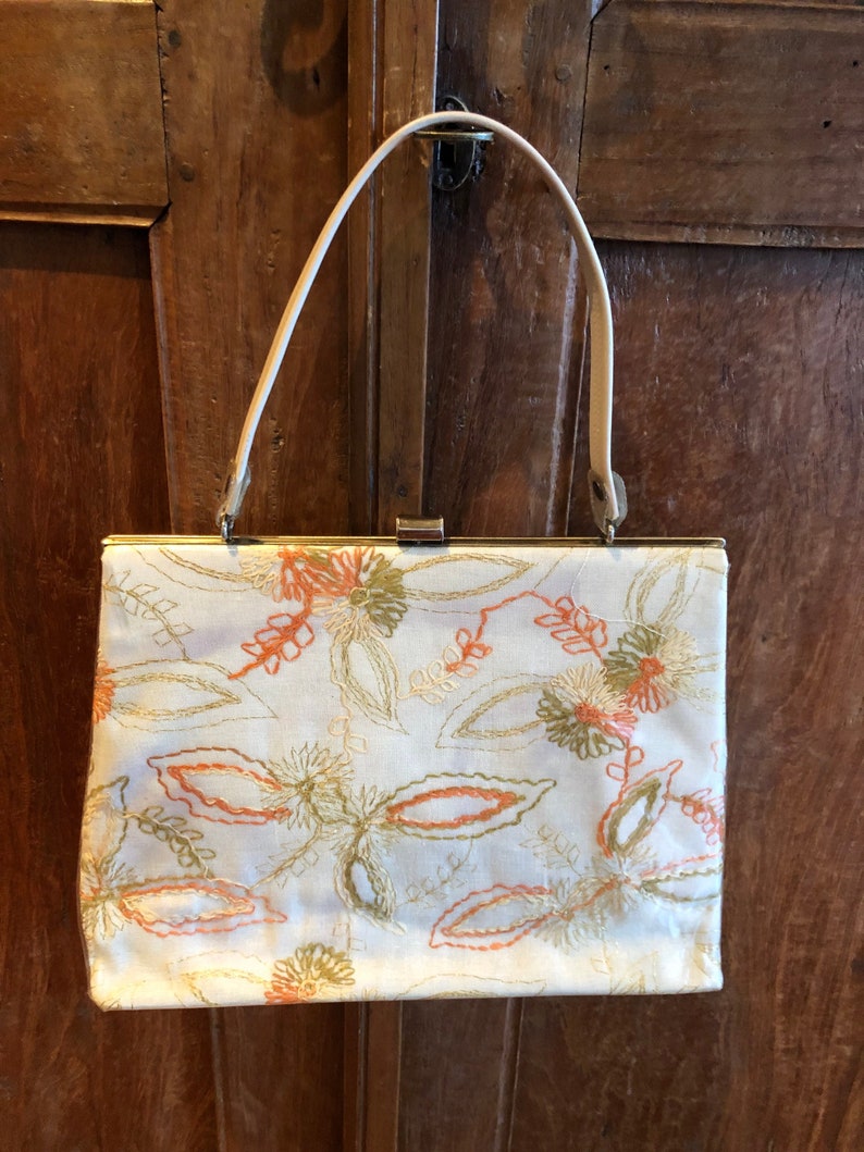 K&ouml;nnte beinhalten: Eine wei&szlig;e Handtasche mit einem floralen Stickmuster in Orange-, Gr&uuml;n- und Goldt&ouml;nen. Die Tasche hat einen Verschluss oben und einen einzigen Trageriemen.