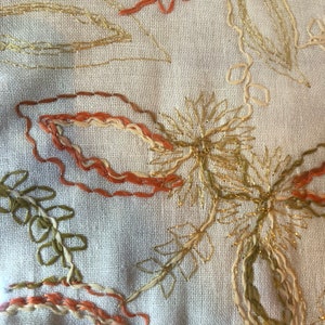 K&ouml;nnte beinhalten: Nahaufnahme eines beigefarbenen Stoffes mit gestickten Blumendesigns in goldener, orangefarbener und gr&uuml;ner Farbe. Die Stickerei weist komplizierte Details und einen zarten, femininen Stil auf.
