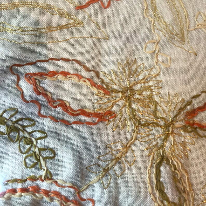 K&ouml;nnte beinhalten: Nahaufnahme eines beigefarbenen Stoffes mit gestickten Blumendesigns in goldener, orangefarbener und gr&uuml;ner Farbe. Die Stickerei weist komplizierte Details und einen zarten, femininen Stil auf.