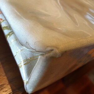 K&ouml;nnte beinhalten: Nahaufnahme der Ecke einer wei&szlig;en Canvas-Tragetasche mit einem transparenten Plastikfenster. Die Tasche hat ein dezentes goldenes Muster.