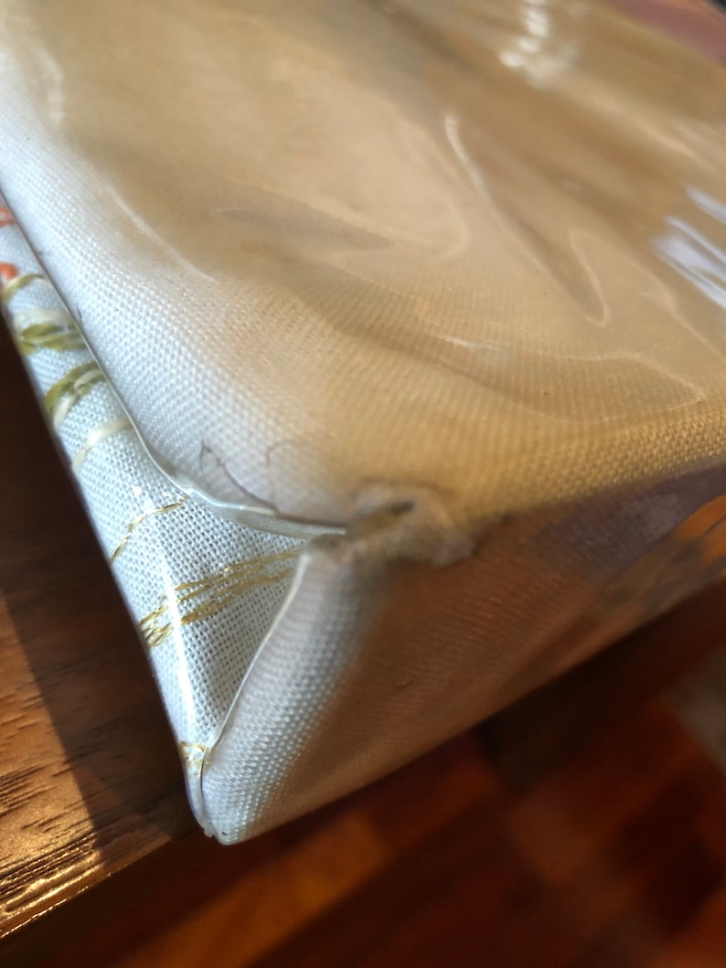 K&ouml;nnte beinhalten: Nahaufnahme der Ecke einer wei&szlig;en Canvas-Tragetasche mit einem transparenten Plastikfenster. Die Tasche hat ein dezentes goldenes Muster.