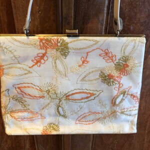 K&ouml;nnte beinhalten: Eine wei&szlig;e Stoff-Clutch mit goldenem Verschluss und einem langen Riemen. Die Tasche ist mit einem floralen Muster in Orange-, Gr&uuml;n- und Cremet&ouml;nen bestickt.