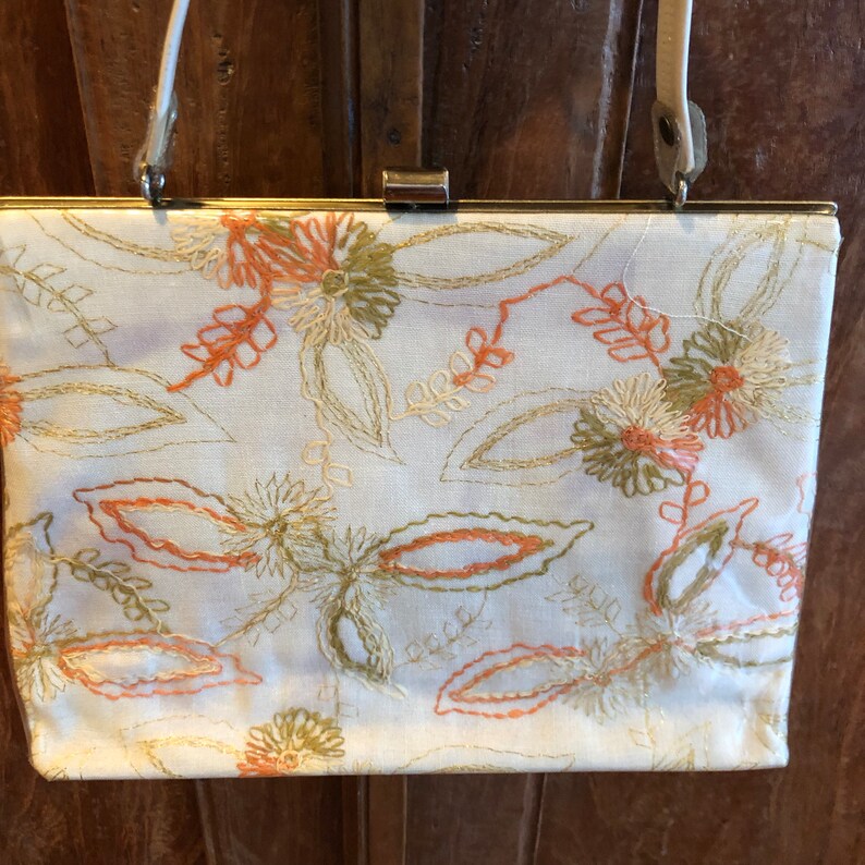 K&ouml;nnte beinhalten: Eine wei&szlig;e Stoff-Clutch mit goldenem Verschluss und einem langen Riemen. Die Tasche ist mit einem floralen Muster in Orange-, Gr&uuml;n- und Cremet&ouml;nen bestickt.