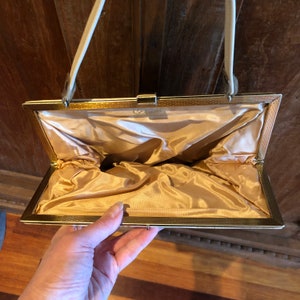 K&ouml;nnte beinhalten: Eine goldfarbene Clutch aus Metall mit einem wei&szlig;en Satin-Futter. Die Clutch hat einen Verschluss oben und einen wei&szlig;en Riemen.