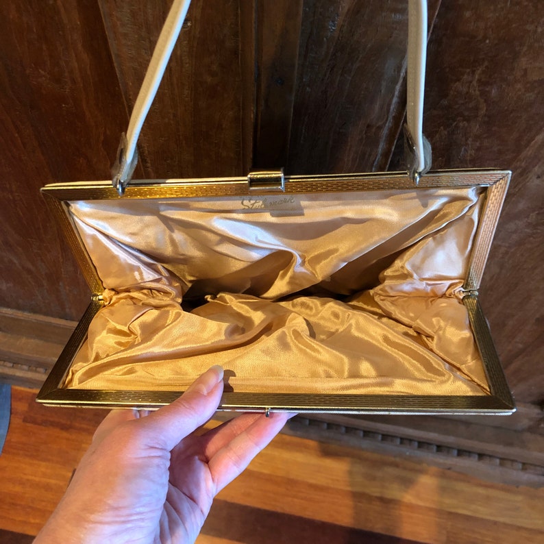 K&ouml;nnte beinhalten: Eine goldfarbene Clutch aus Metall mit einem wei&szlig;en Satin-Futter. Die Clutch hat einen Verschluss oben und einen wei&szlig;en Riemen.