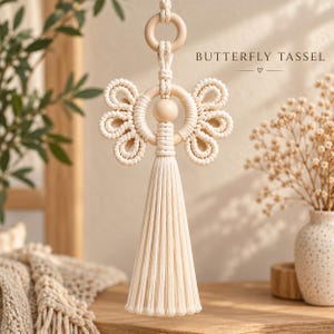 Pode incluir: Um pendente de macramé com uma conta e um anel de madeira central, com cordão de algodão em tom creme atado em forma de borboleta e uma borla longa e reta na parte inferior. O texto BUTTERFLY TASSEL está impresso no canto superior direito.