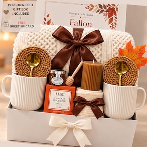 May include: A gift box containing a cream-colored knit blanket with a brown ribbon, two ribbed white mugs with gold spoons on rattan coasters, a jar of honey with a wooden dipper, a box of Lumi Pumpkin Spice Chai tea, and a ribbed brown candle.