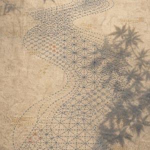 Può includere: Un tessuto beige caratterizzato da un percorso sinuoso di motivi geometrici cuciti in blu. Il tessuto è arricchito da piccoli dettagli ricamati in oro e da un logo rosso nell'angolo in basso a sinistra. Ombre di foglie d'acero sono proiettate.