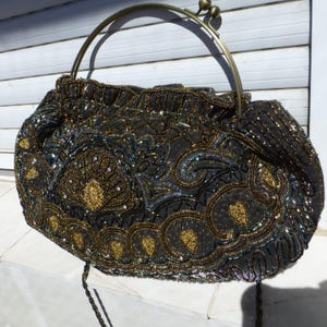 May include: A black handbag featuring intricate beadwork in gold, bronze, and iridescent blue tones. The bag has a rounded shape, a gathered top, and a metallic brass-toned arched handle with a dual-ball clasp closure mechanism at the center.