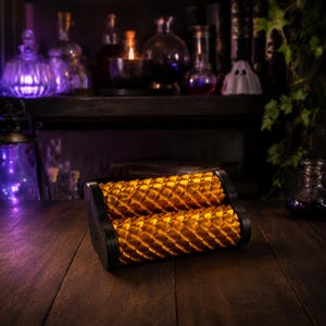 May include: A gold-colored cylindrical roller with a textured scale-like pattern, held between two black circular end caps. The roller features a repeating geometric design across its surface, creating a metallic appearance with light reflections.