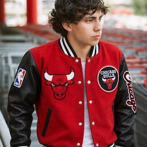 May include: A red varsity jacket with black sleeves, featuring a Chicago Bulls logo on the chest, an NBA patch on the right sleeve, and the word Chicago in script on the left sleeve. The jacket has a white and black striped collar and cuffs.
