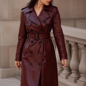 May include: A long, double-breasted trench coat in a deep burgundy colour. The coat features a wide notched collar, shoulder epaulets, long sleeves with buckled cuffs, and a matching waist belt with a rectangular buckle. It has dark buttons.