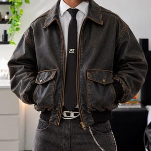 Peut inclure: Un blouson aviateur en cuir marron foncé vieilli avec fermeture éclair, deux poches à rabat et poignets côtelés. Porté sur une chemise blanche à col avec une cravate noire arborant un logo blanc, un jean gris foncé, une boucle de ceinture argentée.