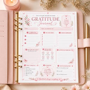 Può includere: Un diario della gratitudine con copertina rosa, anelli dorati e una penna dorata abbinata. La pagina presenta illustrazioni floreali, sezioni per riflessioni quotidiane, suggerimenti numerati, icone dell'umore e il testo Gratitude Journal e Gratitude turns what we have into enough.