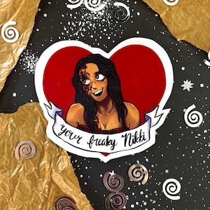 May include: A heart-shaped sticker featuring an illustration of a person with long dark hair and a red mark on their face. A white banner across the bottom contains the black handwritten text "your freaky Nikki" in a cursive script font.