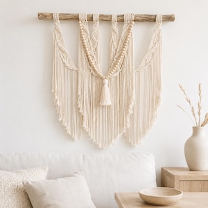 May include: A cream-colored macrame wall hanging suspended from a natural wooden branch. The piece features intricate knotted patterns, a central section adorned with light wooden beads, and a single tassel hanging from the middle point.