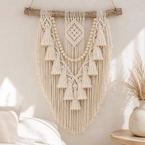 Può includere: Un arazzo in macramè color crema appeso a un ramo di legno orizzontale. Il design presenta intricati motivi a nodi, una trama centrale a forma di diamante, file di perline di legno rotonde e nappe pendenti disposte a forma di V.