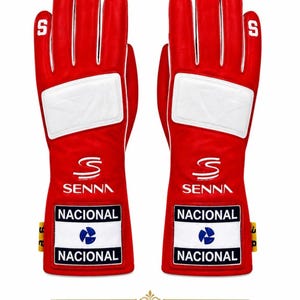 Peut inclure: Une paire de gants de course rouge vif avec des accents blancs, un empiècement rectangulaire blanc sur le dos de la main, et le texte SENNA avec le mot NACIONAL au-dessus et au-dessous d'un logo circulaire bleu sur le poignet. La lettre S est sur les doigts.
