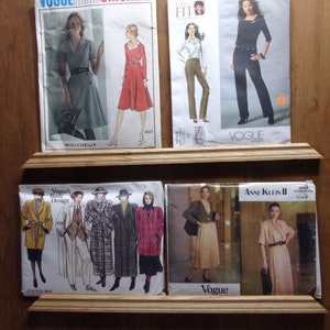 Puede incluir: Una colección de ocho patrones de costura vintage que presentan diferentes diseños para ropa de mujer. Los patrones se muestran en un estante de madera con los nombres de las marcas "Vogue's Basic Design", "Molyneux", "Vogue Patterns", "Anne Klein II" y "Vogue" visibles.