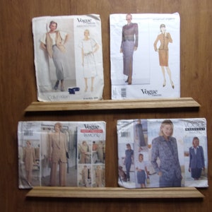 Könnte beinhalten: Vier Vintage-Vogue-Schnittmusterumschläge, die auf Holzregalen ausgestellt sind. Die Muster zeigen Illustrationen von Damenbekleidung, darunter Anzüge, Kleider und Einzelteile. Die Umschläge sind in verschiedenen Farben und Stilen gehalten und zeigen Modetrends.