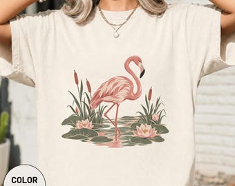 Könnte beinhalten: Ein elfenbeinfarbenes T-Shirt mit einem Grafikdruck eines rosa Flamingos, der in einem Teich steht. Die Illustration zeigt grüne Seerosenblätter, rosa Seerosen und braune Rohrkolben um den Flamingo herum. Das T-Shirt hat kurze Ärmel.