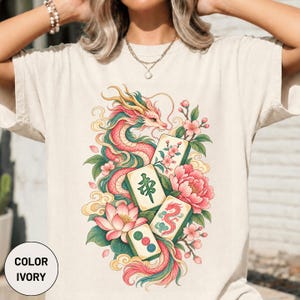 Pode incluir: Uma t-shirt cor de marfim com uma estampa gráfica de um dragão longo e sinuoso em tons de rosa e verde, rodeado por flores de lótus e cerejeiras, intercalada com quatro peças de mahjong exibindo símbolos tradicionais e um ícone de dragão vermelho.
