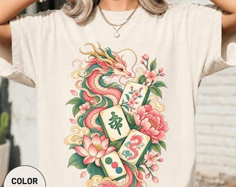 Könnte beinhalten: Ein elfenbeinfarbenes T-Shirt mit einem Grafikdruck eines langen, gewundenen rosa-grünen Drachen, umgeben von blühenden rosa Lotus- und Kirschblüten, durchsetzt mit vier Mahjong-Steinen, die traditionelle Symbole und ein rotes Drachensymbol zeigen.