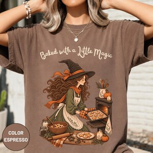 May include: An espresso-coloured t-shirt featuring a graphic of a witch in a pointed hat and apron baking cookies. The text "Baked with a Little Magic" is printed above the illustration, which includes a wooden bowl, rolling pin, and oven.