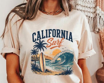 Könnte beinhalten: Ein beiges Kurzarm-T-Shirt mit einem Grafikdruck einer Strandszene mit Surfbrett, Palmen, einer großen Meereswelle und einem Sonnenuntergang. Der Text "CALIFORNIA Surf" ist in dunkelblauer und oranger Schrift über dem Motiv abgebildet.