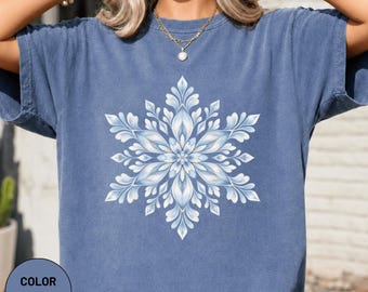 Könnte beinhalten: Ein kurzärmeliges T-Shirt in Jeansblau mit einem großen, symmetrischen, weißen und hellblauen Schneeflocken-Motiv, das mittig auf der Brust aufgedruckt ist. Das T-Shirt hat einen Rundhalsausschnitt und eine lockere Passform mit floralem Muster.