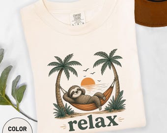 Könnte beinhalten: Ein elfenbeinfarbenes T-Shirt mit einer Grafik eines braunen Faultiers, das in einer Hängematte zwischen zwei Palmen bei Sonnenuntergang ruht. Die Szene zeigt eine orangefarbene Sonne und Vögel. Das Wort "relax" steht in dunkelgrünen Buchstaben darunter.